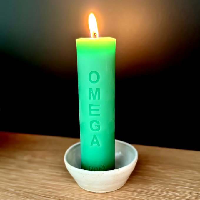 A lit green Omega branded candle sitting in a small white ceramic bowl on a wooden surface against a dark background.