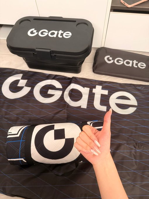 A hand gives a thumbs up next to a collection of Gate.io branded merchandise, including a black carrying case, a mat, a bag, and a rolled item, all featuring the "Gate" logo.