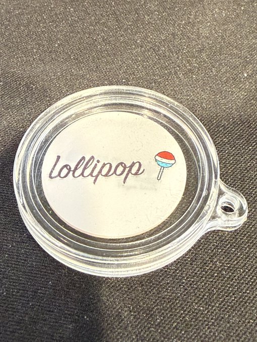 A clear plastic locket charm rests on a dark fabric surface, containing a white insert that reads 'Lollipop' next to a drawing of a red, white, and blue lollipop.