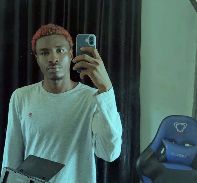 A mirror selfie of a young man with dyed reddish-brown hair wearing a gray long-sleeve shirt, holding a smartphone to take the photo in front of dark curtains, with a blue gaming chair partially visible on the right.