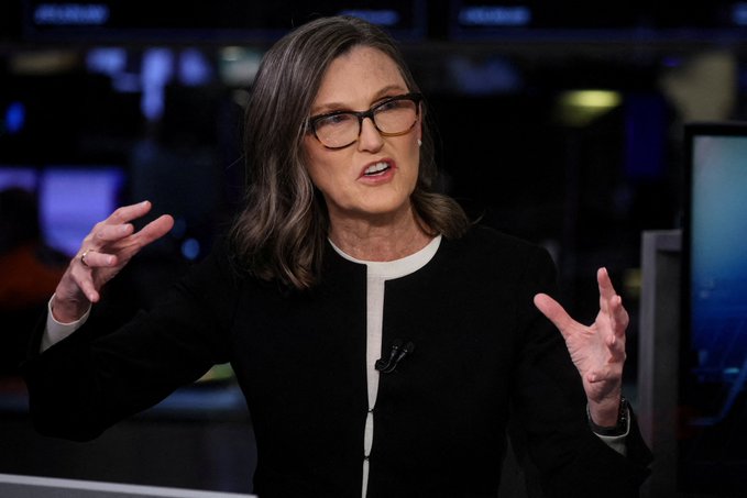 A bespectacled woman with shoulder-length brown hair, wearing a black jacket with white trim, gestures expressively with both hands raised while speaking. The image likely depicts Cathie Wood, CEO of ARK Invest, whose firm recently purchased an additional $37 million in Bitcoin as institutional interest in the cryptocurrency continues to grow despite short-term market volatility.