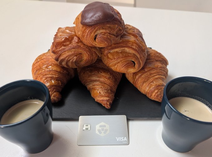 A pyramid of golden croissants, including one topped with chocolate, is arranged on a slate board between two mugs of coffee, with a Crypto.com Visa debit card displayed in front—visually promoting the post's message of earning up to 5% CRO cashback on purchases like this morning coffee break.