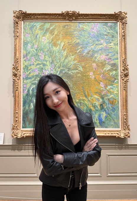 A young woman with long dark hair, wearing a black leather jacket, stands with arms crossed in front of an ornate gold-framed impressionist painting.