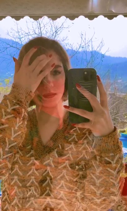 A mirror selfie of a person with red nail polish wearing a beige knit sweater, partially covering their face with one hand while holding a phone with the other, with a view of trees and hills through the window behind them.