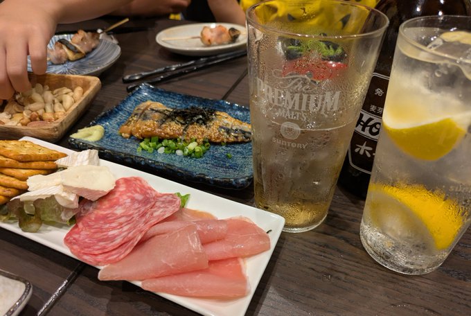A table spread of Japanese food and drinks including sashimi, grilled fish with nori, edamame, peanuts, and glasses of Premium Malt's beer and a lemon highball, with someone's hand reaching in.