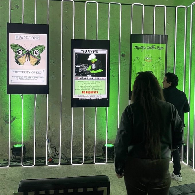 Two attendees at an event look at illuminated green posters displaying 'Butterfly of Kek' and a DJ event flyer mounted on a wall with neon lighting.
