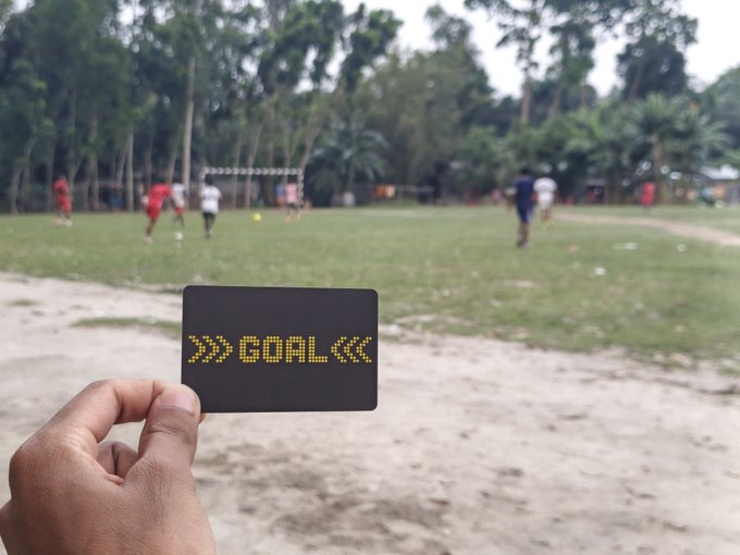 A hand holds up a black card with the pixelated text "» GOAL «" in front of a group of people playing soccer on a grassy field, playing on the double meaning of "goal" as both a soccer term and an objective worth supporting.