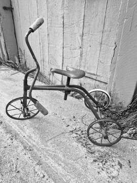 A black and white photograph shows a vintage tricycle parked against a weathered wooden fence.