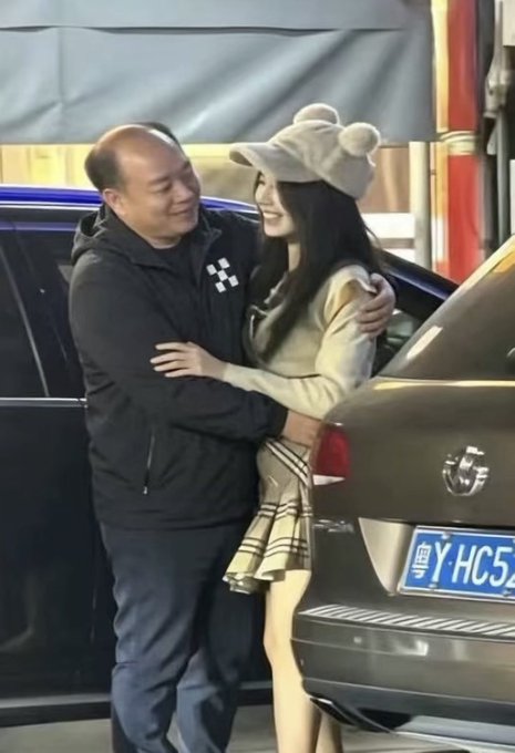 A man in a black hoodie embraces a smiling young woman wearing a bear-eared hat and plaid skirt, standing next to a black car with license plate '粤Y·HC52'.