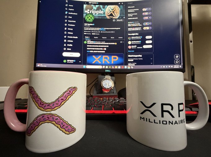 Two coffee mugs sit on a desk in front of a Dell monitor displaying the XRP cryptocurrency's X (Twitter) page—one mug features pink frosted donuts arranged in the shape of the XRP logo, while the other boldly proclaims "XRP MILLIONAIRE." The setup perfectly embodies the post's mantra of coffee and XRP being the essential morning ritual for the crypto faithful.