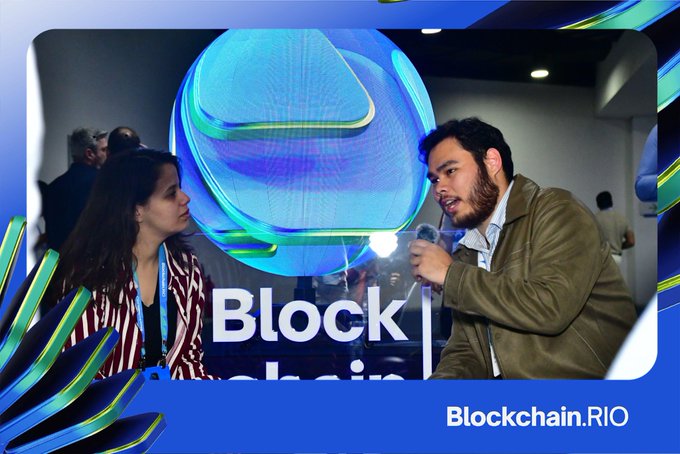 Two people are engaged in conversation at the Blockchain.RIO event, with a large blue and purple digital sculpture or display visible behind them and the "Block" text partially shown on screen. The image reflects the post's context of the speaker attending the event while having their colleague, @brmoniz, represent Nora on the panels rather than speaking themselves.