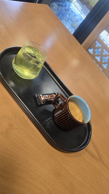 A black oval tray on a wooden counter holds a glass of yellow-green tea, a brown ribbed cup of espresso, and a chocolate bar labeled 'MEGASTAR'.