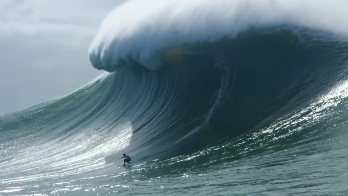 A surfer rides an enormous, towering wave with a frothy white crest, dwarfing the tiny figure on the board against a dramatic ocean backdrop.