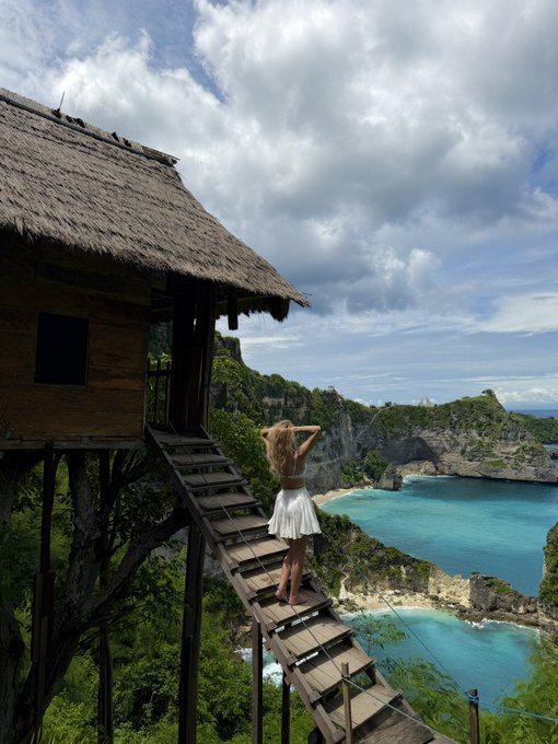 A woman in a white dress stands on a wooden suspension bridge leading from a thatched treehouse, gazing out at a stunning turquoise cove surrounded by dramatic limestone cliffs under a cloudy sky.