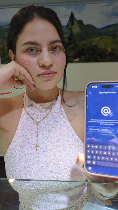 A young woman in a white lacy top rests her chin on her hand while holding a smartphone displaying a Lightning Username (@) setup screen with a keyboard visible.