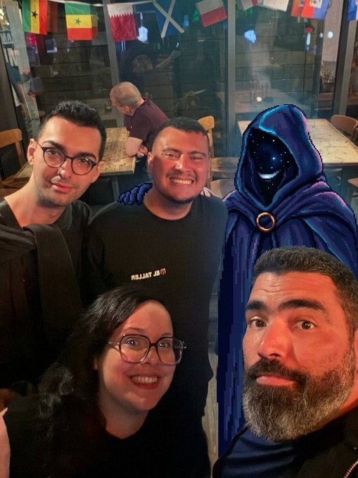 The image features a group of five people posing for a photo, with one person wearing a hooded cape that appears to be a character from a video game or movie. The group consists of four men and one woman, all smiling and enjoying themselves as they pose together in front of a window. In the background, there are flags hanging on the wall, adding a festive atmosphere to the scene. The caption "La Familia dice presente en el Solana Summit de Berlín" suggests that this gathering could be taking place at a convention or event related to video games and gaming culture.