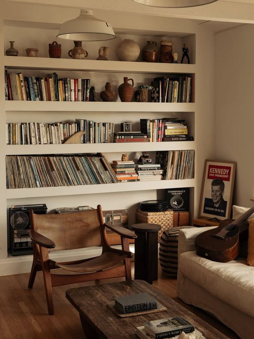 A cozy living room with built-in bookshelves filled with books, vinyl records, and pottery, a brown leather chair, a guitar on a couch, and a Kennedy for President poster.
