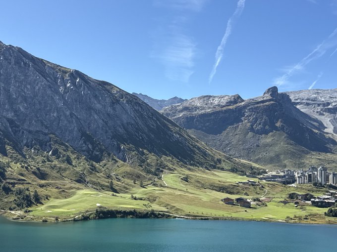 A breathtaking alpine landscape greets the morning, featuring turquoise lake waters nestled among towering mountains and lush green meadows, with what appears to be a ski resort village visible on the distant slopes—perfectly matching the serene "good morning" vibe.
