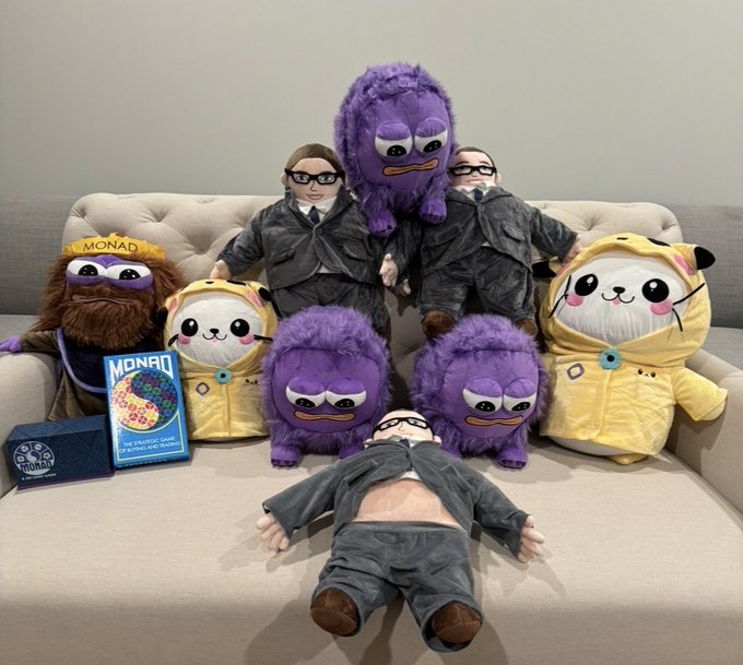 A collection of "Monad" plush toys and figurines in dark suits, hoodies, and goggles are arranged on a couch alongside a "The Official Guide for Monad Developers" booklet, visually echoing the caption's "Monad will win" through a playful army of branded mascots poised for victory.