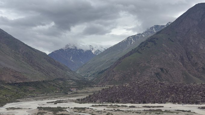A mountain landscape with snow-capped peaks, cloudy skies, and a valley with a winding riverbed in the foreground.