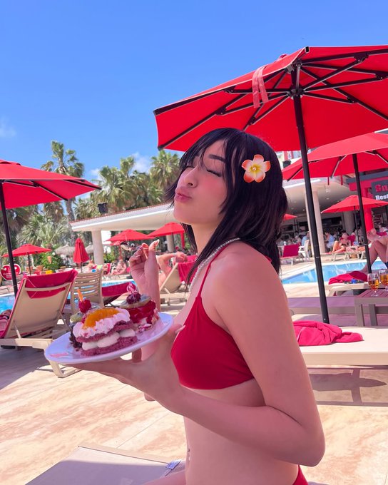 An anime-style character with long dark hair and a flower accessory poses cheerfully while holding a plate of desserts under red umbrellas at a sunny poolside resort.