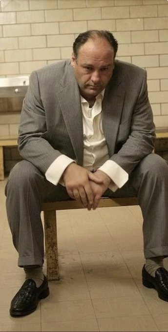 A man in a gray suit sits on a wooden bench in a tiled room, looking contemplative with his hands clasped between his legs.