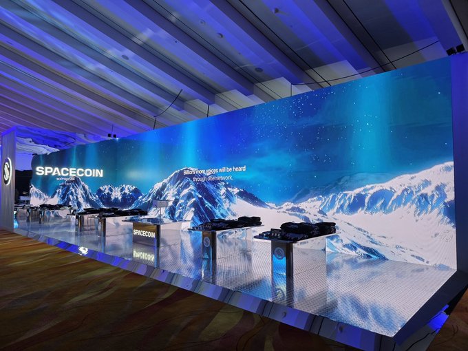 A "SPACECOIN" exhibition booth features illuminated display cases showcasing small satellites, set against a striking backdrop of snowy mountain peaks beneath an aurora-lit starry sky, with the tagline "Blockchain goes will be heard through the cosmos." The satellite models on display visually support the post's mention of bringing satellites to conferences before launching them into orbit.