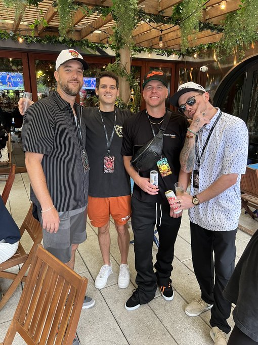 Four men posing together at an outdoor venue, all wearing lanyards, with one holding a beverage can.