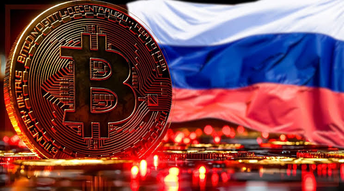 A Bitcoin coin stands prominently in the foreground with the Russian flag waving in the background, visually representing Russia's official enactment of cryptocurrency regulation signed by President Putin. The image symbolically ties the digital asset to the nation's new legal framework governing crypto.

Visible text on the coin includes: "BITCOIN DIGITAL DECENTRALIZED PEER TO PEER."