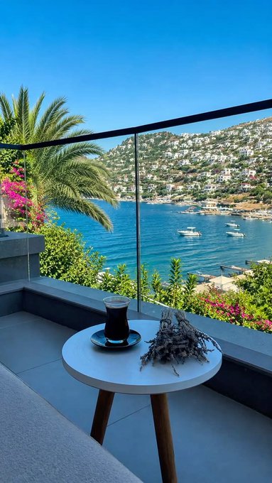 A balcony view overlooking a Mediterranean-style cove with boats and hillside houses, featuring a small table with a Turkish tea glass and dried herbs.