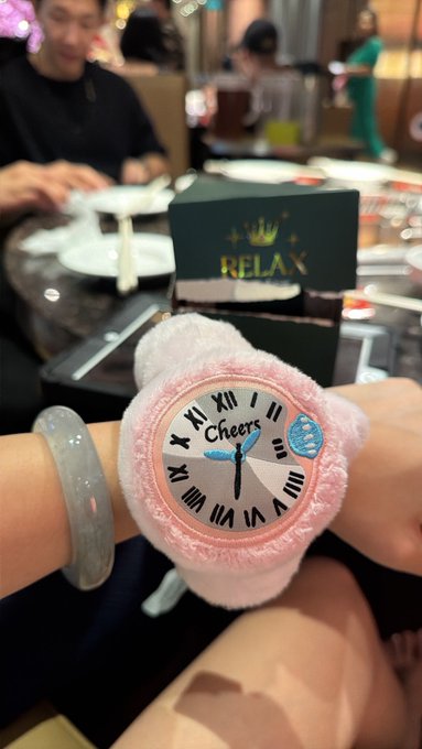 A close-up of a wrist wearing a fuzzy pink watch with the word 'Cheers' on its face and a jade bracelet, taken at what appears to be a restaurant table with a 'RELAX' card in the background.