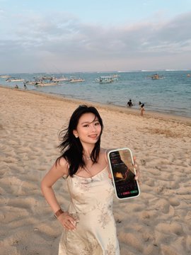 A young woman stands on a beach holding a phone displaying an incoming call, with boats visible on the water behind her.