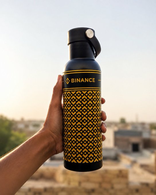 A hand holds up a black and yellow patterned water bottle bearing the 'BINANCE' logo against a hazy cityscape.