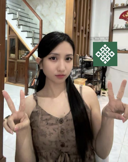 The image features a young woman with long black hair wearing a brown dress, making peace signs with her hands. She is standing next to a staircase, which adds depth to the scene. The background includes a bookshelf filled with various items such as books and vases. The caption of the image reads "#Gritual GM @ritualnet".