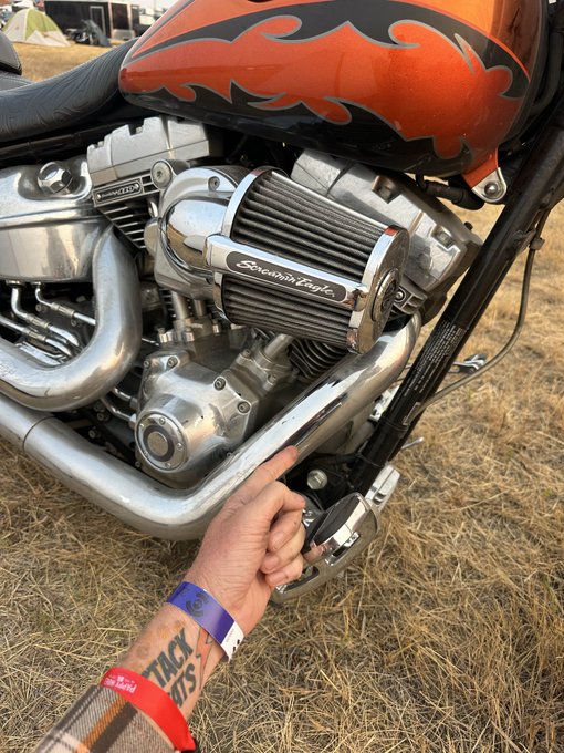 A person's hand with wristbands reaches toward the engine of a custom motorcycle featuring an orange and black flame paint job and a chrome air filter cover labeled 'Screamin' Eagle'.