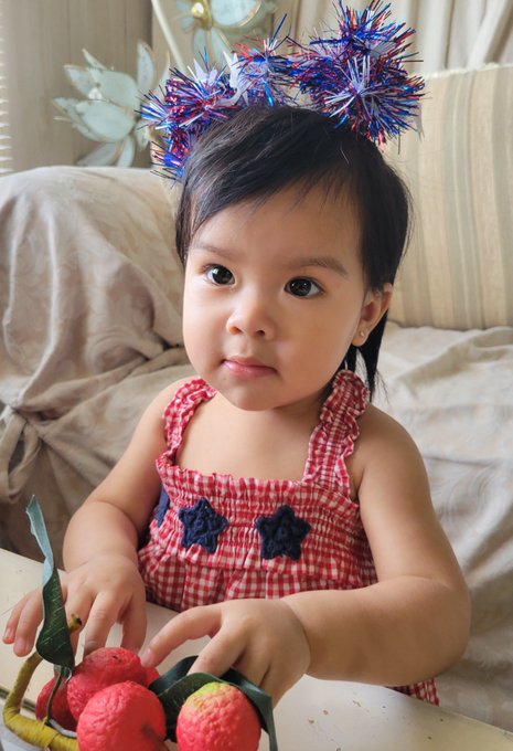 A young child wearing a red and white gingham dress and a festive red, white, and blue tinsel headband sits at a table playing with toy fruits.