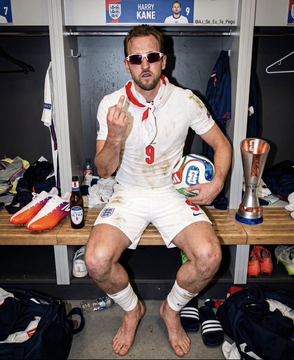A man in a dirty white rugby uniform with a red number '9' sits on a bench in a locker room, holding a rugby ball and a can of beer, with a trophy and soccer cleats visible nearby.