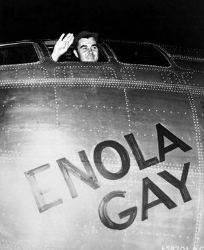 A black-and-white photograph shows a man waving from the cockpit of the B-29 bomber "ENOLA GAY," with the aircraft's name prominently painted in large letters on its metallic fuselage.