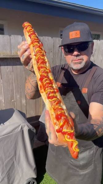 A man wearing a cap, sunglasses, and a brown apron holds up a remarkably long hot dog generously topped with ketchup and mustard in front of a wooden fence.