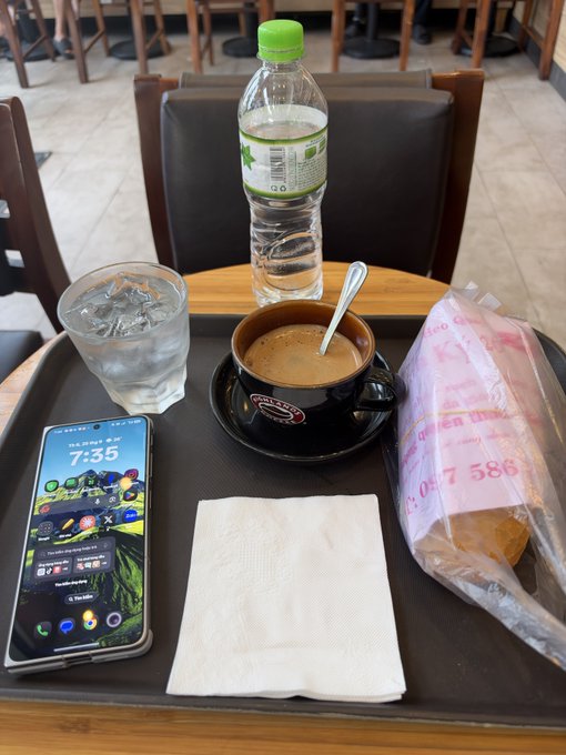 A tray on a cafe table holds a water bottle, a glass of water, an iced coffee, a smartphone displaying the time 7:35, a napkin, and a pink plastic bag with a pastry.