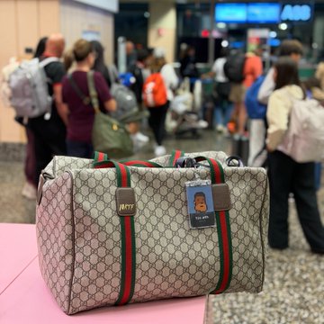 The image shows a large suitcase with a green stripe, sitting on top of a pink table at an airport terminal. The suitcase has a tag that reads "Gm gm @NFC summit I’m coming! Frens see you soon in the sunny Lisbon Punks please don't start brunch without me". This suggests that someone is preparing for a trip and wants to make sure they have all their belongings with them, including their luggage.
