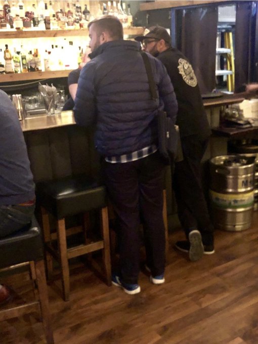 Two men in dark jackets and baseball caps are talking at a wooden bar, with shelves of liquor bottles behind them and a metal beer keg on the floor.