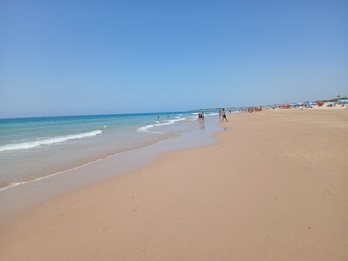 A sunny beach scene with golden sand, gentle blue waves, and people walking along the shoreline under a clear sky.