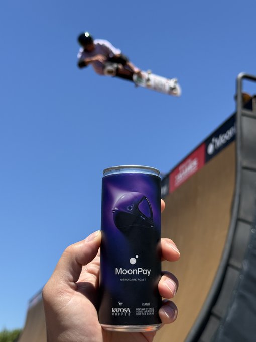 A hand holds up a can of "MoonPay Nitro Dark Roast" coffee in the foreground while an X Games athlete performs an aerial trick on a halfpipe ramp against a clear blue sky in the background. The image pairs MoonPay coffee with the action sports event to match the post's reference to the "MoonPay" and "XGames" duo.