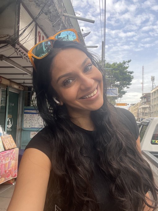 A smiling young woman takes a selfie outdoors on a sunny day, wearing reflective sunglasses perched on her head and a black top, with a street and buildings visible in the background.