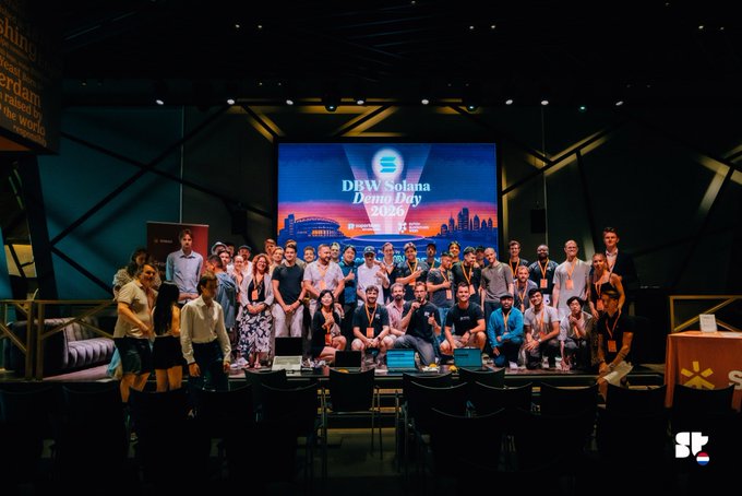 A group photo of participants and organizers poses on stage at the DBW Solana Demo Day 2025, with a large screen displaying "DBW Solana Demo Day 2025" against a cityscape backdrop, capturing the winners and competitors from the Dutch Blockchain Week Summit held at Johan Cruijff ArenA in Amsterdam.