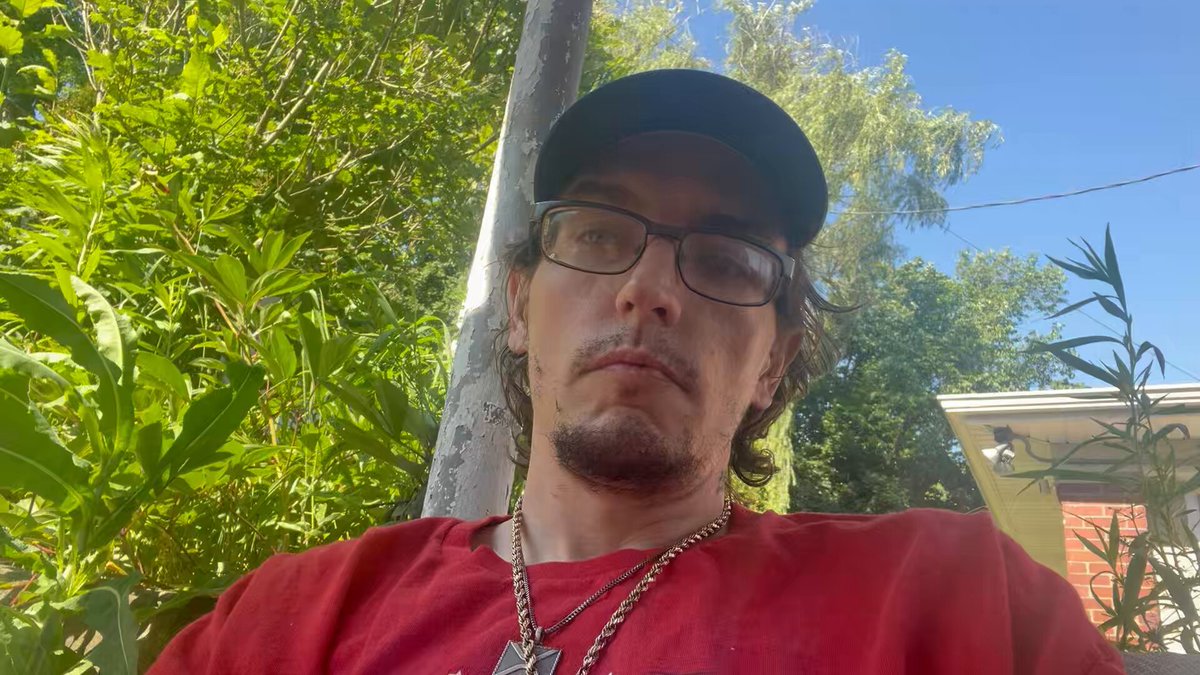 The image features a man wearing glasses, a red shirt, and a black baseball cap sitting outdoors with his eyes closed. The background includes trees and bushes, suggesting that he might be relaxing outside or enjoying some time in nature. There are also several other people visible in the scene, but they do not appear to be the main focus of the image.