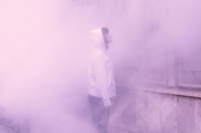 A person in a white hooded jacket and face mask stands enveloped in thick pink and purple smoke next to a concrete wall.