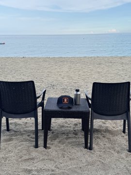 Two dark woven chairs and a small table with items on it sit on a sandy beach facing the ocean under a hazy sky.