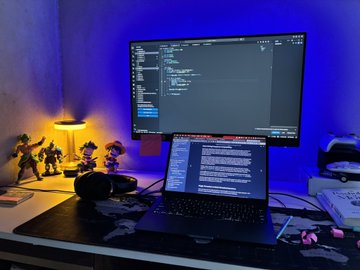 A computer workstation setup featuring an external monitor displaying code and a laptop running terminal/development software, illuminated by blue and orange ambient lighting with small figurines on the desk.